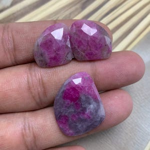 Könnte beinhalten: Drei facettierte, oval geformte Edelsteine in verschiedenen Rosa- und Lilatönen werden gezeigt. Die Edelsteine haben eine polierte, reflektierende Oberfläche mit sichtbaren inneren Mustern. Die Steine werden in einer Hand gehalten, um ihre Farbe und Textur zu präsentieren.