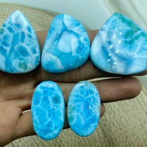Pu&ograve; includere: Cinque pietre preziose di larimar lucide in varie forme. Le pietre mostrano un sorprendente motivo blu e bianco, che ricorda le onde dell'oceano. Le forme includono goccia, triangolare e ovale, mostrando la bellezza naturale del minerale.