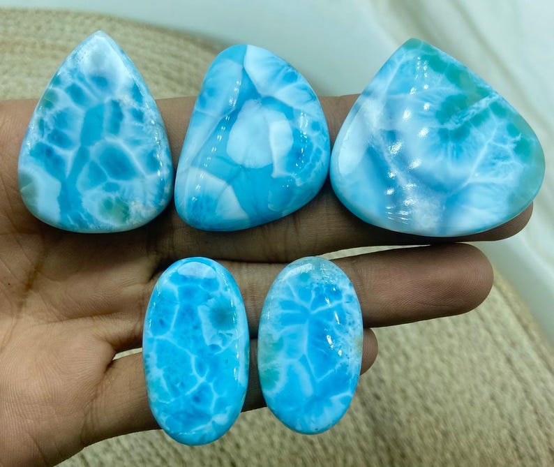 Pu&ograve; includere: Cinque pietre preziose di larimar lucide in varie forme. Le pietre mostrano un sorprendente motivo blu e bianco, che ricorda le onde dell'oceano. Le forme includono goccia, triangolare e ovale, mostrando la bellezza naturale del minerale.