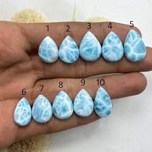 Può includere: Dieci pietre preziose di larimar a forma di goccia, numerate da 1 a 10, presentate su uno sfondo neutro. Le pietre variano in dimensioni e presentano un colore azzurro chiaro con venature bianche, che ricordano le onde dell'oceano. Ogni pietra è lucidata e liscia.