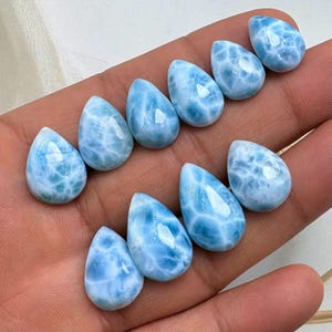 May include: Ten teardrop-shaped larimar gemstones are displayed. The stones are a light blue with white marbling, reminiscent of ocean waves. Each polished stone reflects light. The gemstones are held in a hand. Each stone is approximately 2 cm in length.