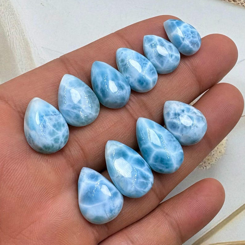 May include: Ten polished larimar gemstones, each in a teardrop shape. The stones are a light blue with white marbling, reminiscent of ocean patterns. The gemstones are held in a hand, showing their size and detail.