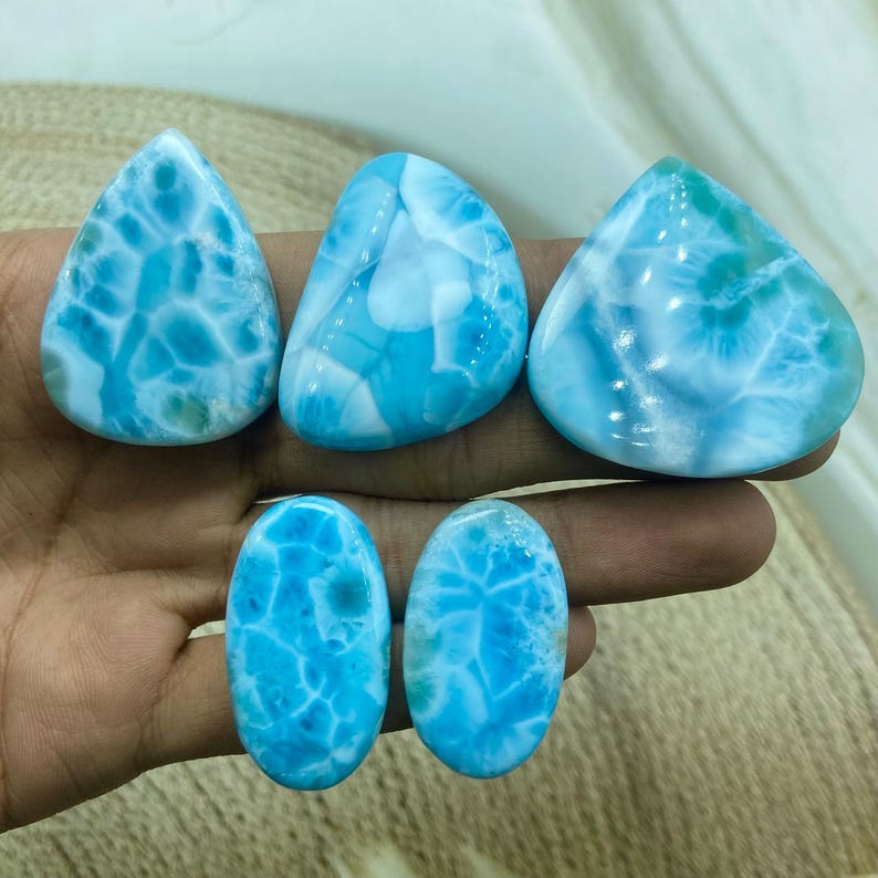 Pu&ograve; includere: Cinque pietre preziose di larimar lucidate in varie forme: a goccia, ovale e irregolare. Le pietre mostrano un sorprendente motivo blu e bianco, che ricorda le onde dell'oceano. Le pietre preziose sono tenute in una mano, mostrando la loro bellezza naturale.