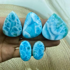 Pu&ograve; includere: Una mano tiene sei pietre preziose di larimar lucide. Le pietre, di varie forme (a goccia, ovali), presentano un sorprendente motivo marmorizzato blu e bianco. Le pietre preziose sono di colore azzurro con venature bianche.