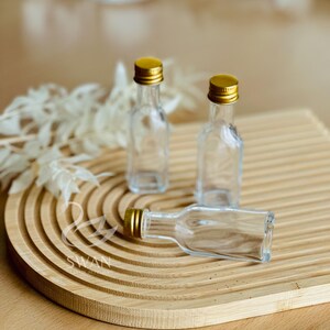 May include: Three small, clear glass bottles with gold-colored caps are arranged on a wooden, ridged serving tray. One bottle is lying on its side. The tray is light brown, and the background is blurred.