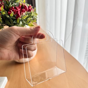 May include: A clear acrylic shopping bag-shaped display holder with a handle. The holder is held in a hand, with a blurred background of a plant with red berries and a window with white curtains.