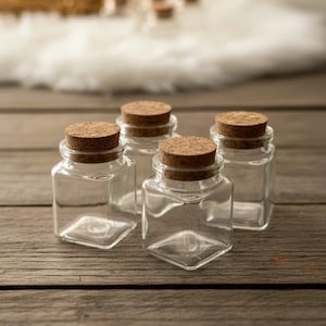 Pode incluir: Quatro pequenos frascos de vidro transparente com corpos quadrados e rolhas de cortiça. Os frascos estão dispostos sobre uma superfície de madeira. As rolhas são de cor castanho claro e os frascos de vidro estão vazios.