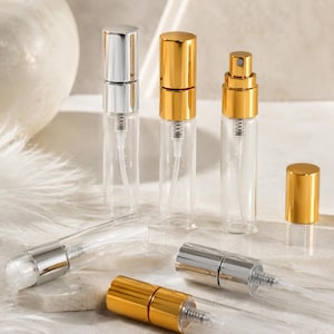 May include: Clear glass perfume atomisers with silver and gold metallic caps. The atomisers are arranged on a light-coloured surface, showcasing their sleek design. Some atomisers are upright, while others are lying down.