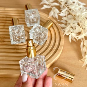 Può includere: Flaconi di profumo in vetro trasparente con vaporizzatori color oro. I flaconi hanno una forma quadrata con un design geometrico strutturato. Sono disposti su un vassoio di legno, con un tappo dorato staccato e fiori bianchi secchi sullo sfondo.