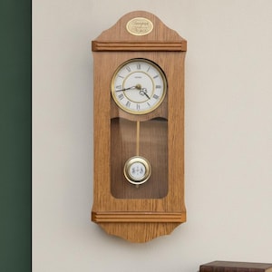 Peut inclure: Une horloge murale en bois avec une finition en chêne clair, dotée d'un cadran blanc circulaire avec des chiffres romains noirs et des aiguilles dorées. Un pendule doré est suspendu derrière une vitre. Une plaque indique Tempus Fugit.