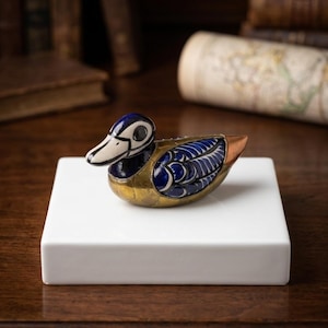 May include: A decorative duck figurine featuring a combination of polished brass and deep blue enamel with white accents. The duck has a smooth, metallic brass chest and tail, with intricate blue and white patterns on its wings and head.