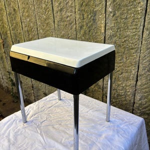 Pode incluir: Uma pequena mesa lateral retangular com tampo branco e corpo preto. A mesa tem quatro pernas cromadas. A mesa está sobre um pano branco. O fundo é uma parede de pedra.