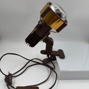 Könnte beinhalten: Ein brauner Klemmstrahler mit einem goldfarbenen zylindrischen Kopf und einer klaren Glaslinse. Die Lampe verfügt über einen dunkelbraunen verstellbaren Arm, einen stabilen Klemmfuß und ein langes braunes Stromkabel mit Zwischenschalter.