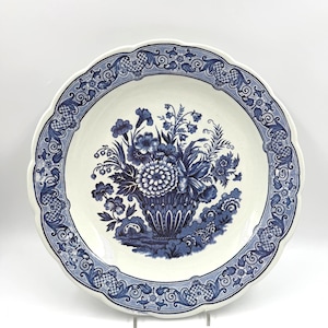 Può includere: Un piatto rotondo in ceramica con bordo smerlato, caratterizzato da una base color crema e un motivo a trasferimento blu scuro. Il centro mostra un bouquet floreale in un cesto, mentre il bordo è decorato con un motivo floreale e a griglia.