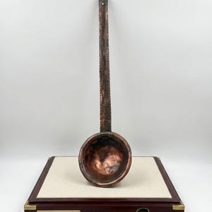 May include: A copper ladle with a long handle and a rounded bowl. The ladle is displayed on a wooden base with the text "CHASING TRADITION: ANCIENT COPPERWARE". A pair of compasses and an open book are also visible.