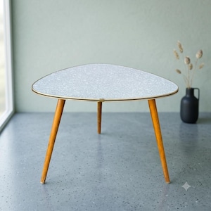 May include: A triangular coffee table with a light gray speckled top and a thin gold-toned metal edge. The table features three tapered wooden legs in a medium brown finish, providing a stable base for the surface. The design is simple and functional.