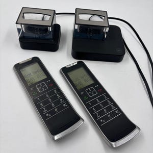 Puede incluir: Dos teléfonos inalámbricos negros con bordes plateados, cada uno con un teclado y una pequeña pantalla que muestra texto. También se ven dos bases de carga con tapas transparentes y bases negras. Los teléfonos y las bases están conectados por cables negros.