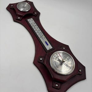 Könnte beinhalten: Wetterstation aus weinrotem Leder mit drei silberfarbenen Messgeräten. Das obere Messgerät ist ein Kompass, das mittlere ein Thermometer und das untere ein Barometer. Das Leder ist genäht und genietet.