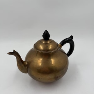 Może przedstawiać: Zabytkowy mosiężny czajnik o zaokrąglonym korpusie, zakrzywionej wylewce i czarnym uchwycie. Pokrywka zwieńczona jest czarnym guzikiem. Czajnik ma ciepłe, metaliczne wykończenie i jest umieszczony na gładkim, białym tle.