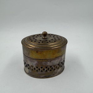 Peut inclure: Petit récipient rond en métal antique avec couvercle. Le couvercle et la partie supérieure sont dorés, tandis que la partie inférieure est argentée avec des motifs découpés décoratifs. Un petit bouton est posé sur le couvercle. Le récipient est probablement utilisé pour le stockage.