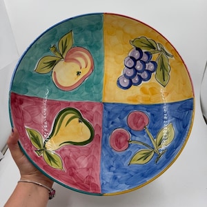 Könnte beinhalten: Eine runde Keramikschale, die in vier Quadranten unterteilt ist, von denen jeder eine handgemalte Obstillustration zeigt. Die Abschnitte zeigen einen gelben Apfel auf Türkis, violette Trauben auf Gelb, eine gelbe Birne auf Magenta und rote Kirschen auf Blau.