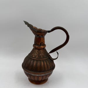 Op de afbeelding: Een koperkleurige metalen kan met een bolvormig lichaam met verticale ribbels en een geschulpte rand. De kan heeft een smalle hals met een decoratieve band en een gebogen handvat aan de zijkant, afgewerkt met een donkere patina.