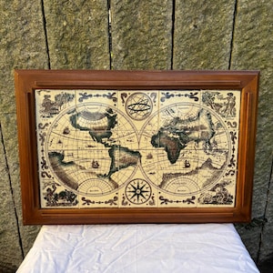 Könnte beinhalten: Eine gerahmte Vintage-Weltkarte mit detailliertem Design. Die Karte zeigt Kontinente in Braun- und Grüntönen, mit dekorativen Illustrationen und einer Windrose. Der Rahmen ist aus dunklem Holz.