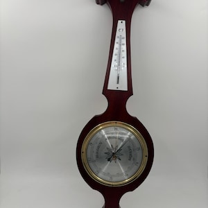 May include: A vintage wooden barometer with a thermometer. The barometer has a circular face with a gold-coloured rim and a silver dial. The thermometer displays both Celsius and Fahrenheit scales. The wood is a dark reddish-brown colour.