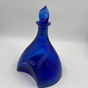 Puede incluir: Una jarra de vidrio azul cobalto con un diseño curvo único. La jarra presenta un tapón en forma de lágrima. El vidrio tiene un acabado brillante, que refleja la luz. Esta pieza decorativa es adecuada para servir bebidas.