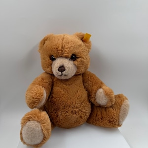Puede incluir: Un oso de peluche marrón con hocico y almohadillas de las patas de color marrón claro. El peluche tiene ojos oscuros y una pequeña etiqueta amarilla en la oreja. El oso está sentado con las piernas separadas.
