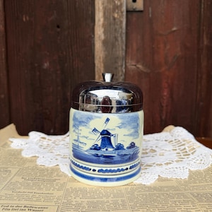 Può includere: Vaso in ceramica blu e bianca con coperchio argentato, con un disegno di mulino a vento. Il vaso cilindrico è su un giornale con un centrino di pizzo. Estetica vintage e decorativa.