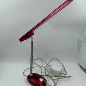 May include: A red desk lamp with a rectangular light head and a silver adjustable arm. The lamp has a red base with a white power button and a white power adapter. The lamp is designed for desk use.