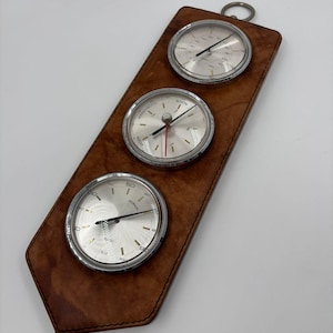 Puede incluir: Una estación meteorológica de cuero marrón con tres medidores plateados. Los medidores tienen agujas negras y bordes plateados. La parte trasera de cuero tiene un anillo de metal en la parte superior para colgar.