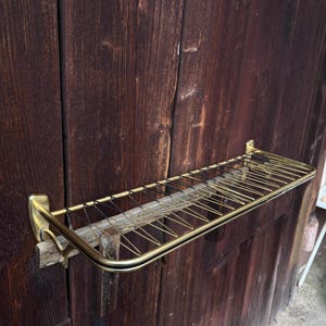 May include: A vintage brass-toned wall-mounted shelf with a wire rack design. The shelf is attached to a weathered wooden surface, showcasing a classic, functional design. The shelf is approximately 24 inches long.