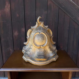 May include: A hand-carved wooden decorative element featuring a bell-like shape with a central circular medallion, scalloped edges, and a crescent moon motif at the top. The wood displays a natural grain with dark brown staining in the recessed areas.