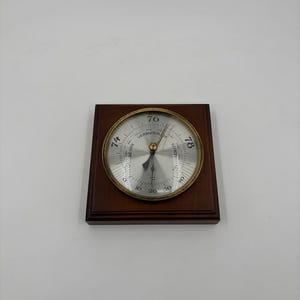 May include: A vintage barometer with a brass bezel and a silver face, set on a dark brown wooden base. The face shows numbers from 10 to 80, with the word "VERANDERL" visible.