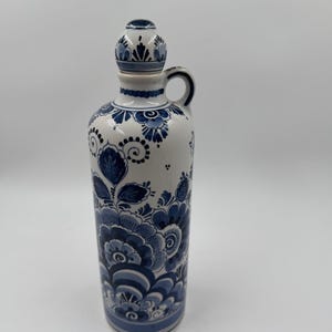 Może przedstawiać: Biała ceramiczna butelka z niebieskim kwiatowym wzorem. Butelka ma uchwyt i korek. Wzór kwiatowy obejmuje stylizowane kwiaty i liście w różnych odcieniach niebieskiego. Butelka ma około 25 cm wysokości.