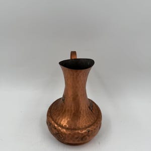 May include: A small copper jug with a hammered texture and a rounded base. The metal surface displays a warm, reddish-brown metallic finish with visible indentations across the body. It features a narrow neck and a small handle at the top.