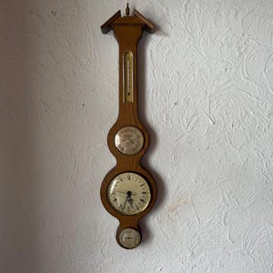 May include: A vertical wooden wall-mounted weather station featuring a thermometer at the top, a barometer in the center, a clock with Roman numerals below it, and a small hygrometer at the bottom, all encased in a light brown wood frame.