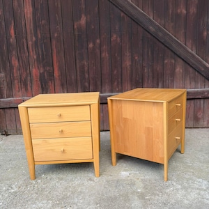 Könnte beinhalten: Zwei Nachttische aus hellem Holz, jeweils mit drei übereinanderliegenden Schubladen und kleinen, runden Holzknöpfen. Die Nachttische sind mit vier konisch zulaufenden Beinen konstruiert und haben eine glatte Oberfläche mit natürlicher Holzmaserung.