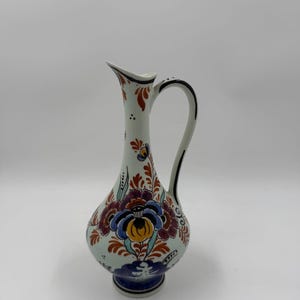 Puede incluir: Una jarra de cerámica blanca con cuello delgado y asa curva. La jarra está adornada con un diseño floral en tonos azules, rojos, amarillos y verdes. La base es azul oscuro y el asa es blanca con ribete negro.