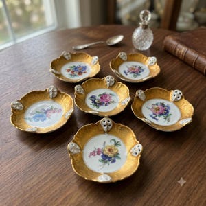 Könnte beinhalten: Ein Set aus sechs kleinen Porzellanschalen mit gewelltem Rand, Goldverzierungen und weißen Zentren mit bunten Blumensträußen. Jede Schale hat drei kleine, weiße, durchbrochene Zierlaschen am äußeren Rand der goldenen Umrandung.