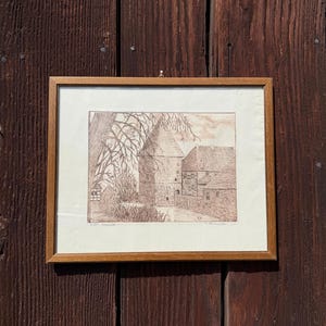 May include: A framed sepia print depicting a building with a tower and a tree. The artwork is in a wooden frame, set against a dark brown wooden background. The print appears to be an architectural drawing or etching.