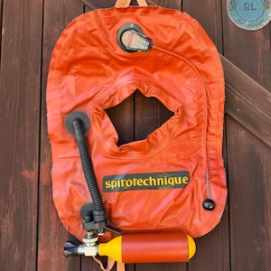 May include: An orange buoyancy compensator vest featuring the text spirotechnique in yellow letters on a black background. The vest includes a black corrugated inflation hose, a small yellow cylinder, and a clear plastic valve component at the top.