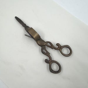 May include: A pair of antique-style brass wick trimmers featuring ornate, scroll-work handles with two finger loops and a spring-loaded mechanism. The metal has a darkened, aged patina, and the blades are positioned at an angle to the handle.