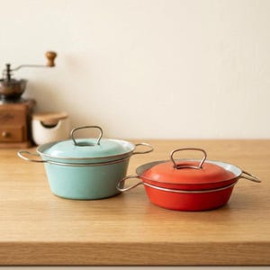 Op de afbeelding: Twee ronde metalen kookpannen met deksels en zijhandgrepen. De ene pan is lichtblauw en de andere is oranje. Beide pannen zijn voorzien van een metalen rand en een bijpassende metalen handgreep op het deksel. Ze staan naast elkaar op een vlak.