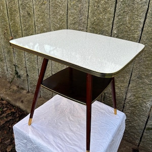 以下が含まれることがあります： 白の模様入りの天板と濃い茶色の棚が付いた、ヴィンテージのサイドテーブル。 テーブルは、先細りの赤茶色の脚に金色のチップが付いています。 テーブルの寸法は約61cmの長さ、41cmの幅、51cmの高さです。