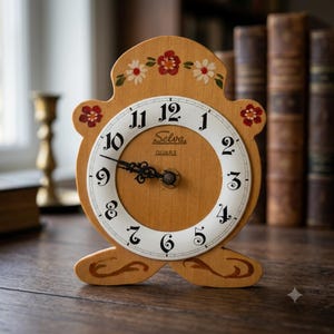 Puede incluir: Un reloj de sobremesa de madera marrón claro con parte superior y base redondeadas. La esfera es blanca con números arábigos negros y el texto Selva Quarz. El marco de madera presenta motivos florales pintados a mano en rojo y blanco.
