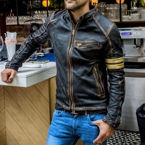 以下が含まれることがあります： ヴィンテージ感のある、黒い革のジャケット。フロントはジッパー開閉、複数のポケット、左袖にはゴールドのストライプが入っています。青いジーンズと合わせています。
