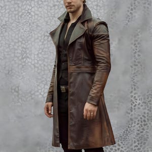 May include: A brown leather trench coat with a notched collar and a belt. The coat has a long, flowing design and is worn over a black vest and shirt. The coat's color is a mix of brown and bronze, with a slight sheen.